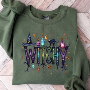May include: An olive green sweatshirt with the word "WITCHY" in a colorful, whimsical font. The design includes a witch's hat, potion bottles, a black cat, and stars. The sweatshirt is laid flat on a wooden surface.