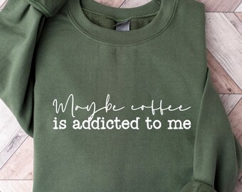 May include: An olive green sweatshirt with the white text "Maybe coffee is addicted to me". The long-sleeved crew neck top is made of a soft material and is laid flat on a wooden surface.