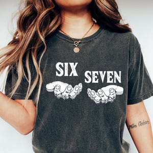 May include: Dark gray t-shirt with the words "SIX SEVEN" in white block letters above two white hand illustrations. The shirt is paired with blue jeans and white slip-on shoes. The overall style is casual and relaxed.