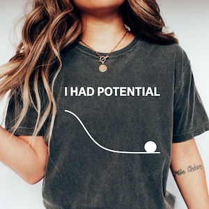 May include: Dark gray t-shirt with the white text "I HAD POTENTIAL" above a graphic of a ball rolling down a curved line. The shirt is paired with blue jeans and white sneakers.