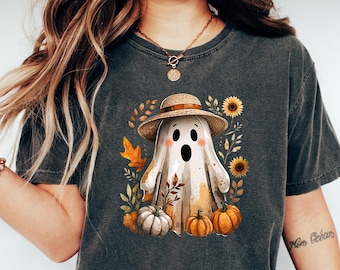 May include: Dark gray t-shirt featuring a whimsical ghost wearing a straw hat, surrounded by pumpkins, sunflowers, and fall foliage. The ghost has black eyes and a smiling mouth. The shirt is paired with blue jeans and white sneakers.