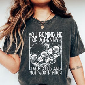 May include: Dark gray t-shirt with the text "YOU REMIND ME OF A PENNY TWO-FACED AND NOT WORTH MUCH" and a graphic of two skulls. The shirt is worn with blue jeans and white shoes.