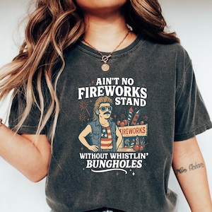 May include: Dark gray t-shirt with the text "Ain't No Fireworks Stand Without Whistlin' Bungholes." The graphic features a cartoon man with sunglasses and a mullet, standing in front of a fireworks stand. The shirt is paired with blue jeans.