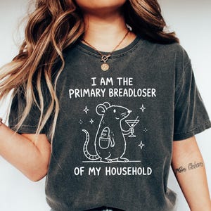 May include: Dark grey t-shirt featuring the text "I AM THE PRIMARY BREADLOSER OF MY HOUSEHOLD" and a cartoon rat with a cocktail and a baguette. A casual, everyday wear t-shirt.