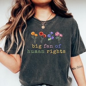 May include: Dark gray t-shirt with the text "big fan of human rights" in rainbow colors. The shirt features a floral design above the text. The model is wearing blue jeans and white sneakers.
