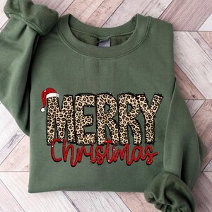 May include: An olive green sweatshirt with the words "MERRY Christmas" in a leopard print design and red script. A Santa hat is on top of the word "MERRY". The sweatshirt is folded on a light brown and white wooden surface.