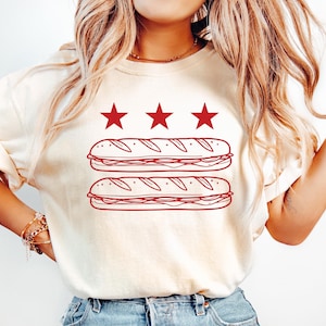 May include: Cream-colored t-shirt featuring a red graphic of two sub sandwiches and three red stars. The design is centered on the front of the shirt. The shirt is paired with light blue denim jeans.