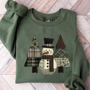 May include: Green crewneck sweatshirt with a snowman and two Christmas trees appliqué. The snowman has a black top hat, scarf, and button details. The trees and snowman are made of patchwork fabric. The sweatshirt has ribbed cuffs and hem.