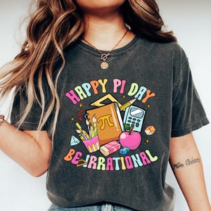 May include: Dark gray t-shirt with a colorful graphic that says "Happy Pi Day Be Irrational." The graphic includes a book, apple, calculator, pencils, and other school supplies. The shirt is paired with blue jeans and white shoes.