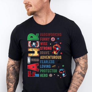 May include: Black t-shirt with the word "FATHER" in colorful block letters. The shirt lists qualities like "hardworking," "funny," and "hero" with superhero-themed graphics. A man is wearing the shirt.