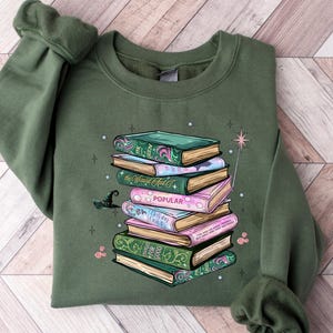 May include: Olive green sweatshirt featuring a stack of colorful books with a witch hat and a magic wand. The books have decorative covers and the word "POPULAR" is visible on one. The design is whimsical and book-themed.