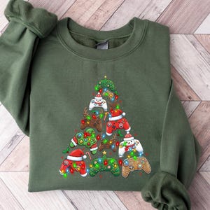 May include: An olive green sweatshirt with a Christmas tree design composed of colourful video game controllers. The controllers are adorned with Santa hats and fairy lights, creating a festive holiday theme.
