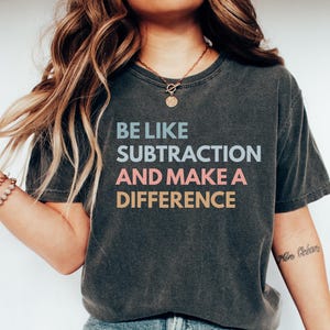Peut inclure: T-shirt gris foncé avec l'inscription "BE LIKE SUBTRACTION AND MAKE A DIFFERENCE" en bleu, rose et jaune. Le t-shirt est porté avec un jean bleu et des chaussures blanches. Un collier et des bracelets sont également visibles.