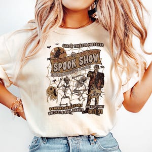 May include: A cream-coloured t-shirt with a Halloween-themed graphic. The design features the words "SPOOK SHOW" and illustrations of skeletons, a zombie, and a ghost. The shirt also has the text "OCT 31".