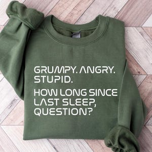 May include: An olive green sweatshirt with the text "GRUMPY. ANGRY. STUPID. HOW LONG SINCE LAST SLEEP, QUESTION?" in white. The sweatshirt is folded on a light brown wooden surface.