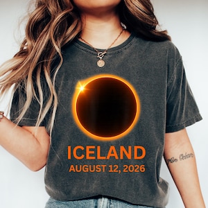 Peut inclure: T-shirt gris foncé avec un motif d'éclipse solaire, avec un contour orange vif et un centre noir. Le mot "ICELAND" est imprimé en orange, ainsi que la date "12 AOÛT 2026". Le t-shirt est porté avec un jean bleu et des baskets blanches.