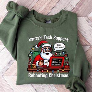 May include: An olive green sweatshirt featuring the text "Santa's Tech Support" and a pixelated Santa Claus graphic. Santa is at a computer with a "Critical Error" message. The text "Rebooting Christmas" is below the graphic.