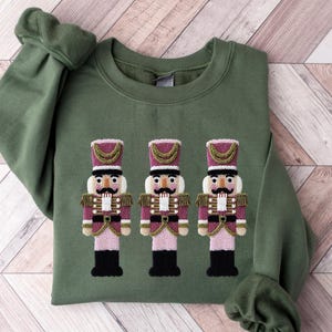 May include: An olive green sweatshirt with an embroidered design of three toy nutcracker soldiers. The nutcrackers have pink and purple uniforms with gold accents and black boots. The sweatshirt has long sleeves and a crew neck.