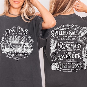 May include: A charcoal gray short-sleeve t-shirt featuring white graphic prints. The front displays a mortar and pestle with the text "Owens Sisters Apothecary Est. 1995". The back features text "Always throw spilled salt over your left shoulder, keep rosemary by your garden gate, plant lavender for luck, and fall in love whenever you can."