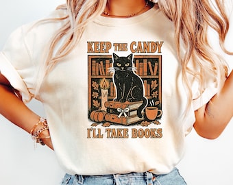 May include: Cream-colored t-shirt featuring a graphic of a black cat sitting on books in front of a bookshelf. The text reads "Keep the Candy, I'll Take Books" in a vintage style. The design includes a candle, pumpkin, and a cup of coffee.