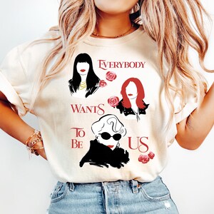 May include: Cream-colored t-shirt featuring a graphic design with three stylized female portraits and the text "EVERYBODY WANTS TO BE US" in red. The design includes roses. The shirt is paired with light blue jeans.