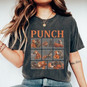May include: Dark gray t-shirt with the word "PUNCH" in orange block letters. The shirt features a grid of nine squares, each with a different illustration of cartoon monkeys. The model is wearing blue jeans and white sneakers.