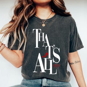 May include: Dark gray t-shirt with the text "THAT'S ALL" in white, with a red high heel shoe graphic. The shirt is paired with blue jeans and white sneakers. The overall style is casual and relaxed.