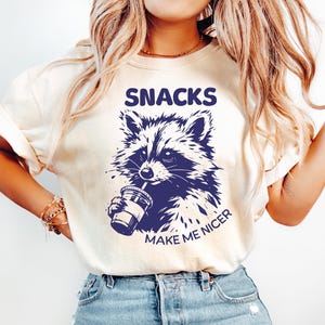 May include: A cream-coloured t-shirt featuring a navy blue raccoon illustration drinking from a cup. The words "SNACKS" and "MAKE ME NICER" are printed above and below the raccoon. The shirt is worn with light blue jeans.