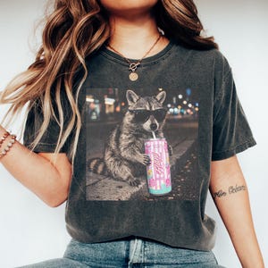May include: Dark gray t-shirt featuring a graphic of a raccoon wearing sunglasses and holding a pink can with the word "Alani" on it. The raccoon is sitting on a sidewalk with a blurred city background. The shirt is paired with blue jeans.