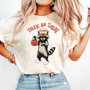 Może przedstawiać: Kremowy t-shirt z grafiką szopa trzymającego lampion z dyni i noszącego kapelusz. Tekst "Trick or Treat" jest wygięty nad szopem. Szop nosi białe buty. Koszulka jest połączona z jasnoniebieskimi dżinsami.