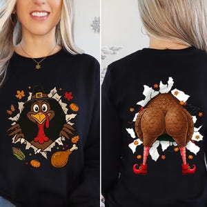 May include: Black long-sleeve sweatshirt with a Thanksgiving turkey graphic. The front features a turkey wearing a pilgrim hat bursting through a torn hole. The back shows the turkey's rear and legs with red shoes bursting through a similar hole.