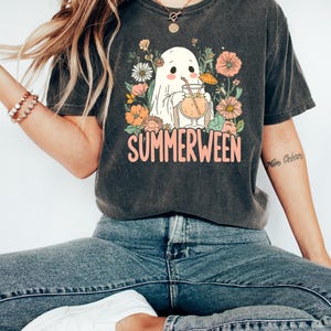 Op de afbeelding: Donkergrijs T-shirt met een afbeelding van een spook dat een cocktail drinkt, omringd door bloemen. Het woord "SUMMERWEEN" is in roze gedrukt. Het shirt wordt gedragen met een blauwe jeans en witte sneakers.