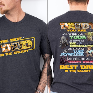 Puede incluir: Camiseta gris oscuro con el texto "THE BEST DAD IN THE GALAXY" en amarillo y naranja. La parte trasera de la camiseta tiene el texto "DADDY" y varios personajes de Star Wars, incluyendo a Yoda y Darth Vader.