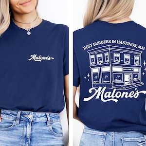 May include: A navy blue t-shirt featuring the white text "Malone's" on the front chest. The back displays a white line-art illustration of a building with the text "BEST BURGERS IN HASTINGS, MA!" and the word "Malone's" in a large script font.