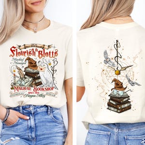May include: Cream-colored t-shirt with a front graphic of a magical bookshop scene, including a stack of books, a sorting hat, and an owl. The back features a similar design with a golden snitch and flying owls. Text includes "Flourish & Blotts" and "Magical Bookshop."