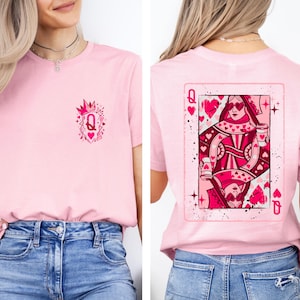 May include: Pink t-shirt featuring a Queen of Hearts design. The front of the shirt has a small graphic with a crown and the letter "Q". The back displays a playing card with a Queen of Hearts illustration. The shirt is paired with blue jeans.