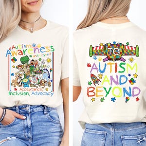 K&ouml;nnte beinhalten: Cremefarbenes T-Shirt mit farbenfrohem Text und Grafiken zur F&ouml;rderung des Autismus-Bewusstseins. Die Vorderseite zeigt ein Cartoon-Design mit den Worten "Autismus-Bewusstsein und dar&uuml;ber hinaus, Akzeptanz, Inklusion, Advocacy". Die R&uuml;ckseite zeigt "Autismus und dar&uuml;ber hinaus" mit Puzzleteilen und Sternen.
