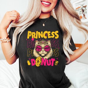May include: Black t-shirt with a graphic design featuring a cat wearing sunglasses and a crown, with the words "PRINCESS" and "DONUT" in yellow and pink. The cat graphic includes the text "THIS OUT RAGE".