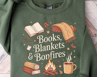 May include: Green sweatshirt with the text "Books, Blankets & Bonfires" and illustrations of a book, blanket, campfire, and mug. The design features fall-themed elements in shades of brown, orange, and white. The sweatshirt has long sleeves.