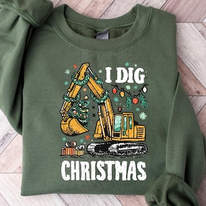 Peut inclure: Sweat-shirt vert avec le texte "I DIG CHRISTMAS" et un dessin de pelle mécanique jaune décorée d'un sapin de Noël et de lumières. Des cadeaux sont au bas de la pelle.