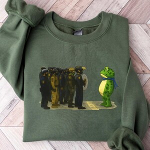 May include: An olive green sweatshirt featuring a graphic of a frog facing a group of figures in uniform. The frog is green with yellow spots and a blue scarf. The figures are in dark uniforms.