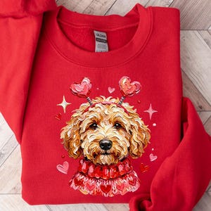 May include: A red sweatshirt featuring a detailed illustration of a golden doodle dog wearing a heart-themed headband and sweater. The design includes hearts and stars, creating a festive and loving theme.