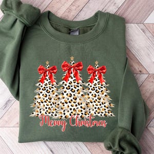 May include: An olive green sweatshirt with a Christmas-themed design. The design features three leopard print Christmas trees, each with a red bow and star. The words "Merry Christmas" are written in red script below the trees.