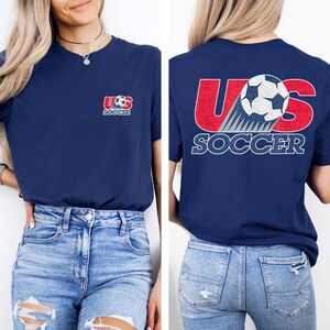 Puede incluir: Una camiseta azul marino con un peque&ntilde;o gr&aacute;fico en el pecho izquierdo que dice US SOCCER con un bal&oacute;n de f&uacute;tbol. La parte posterior de la camiseta muestra un gr&aacute;fico grande con el texto rojo US, un bal&oacute;n y el texto blanco SOCCER debajo.