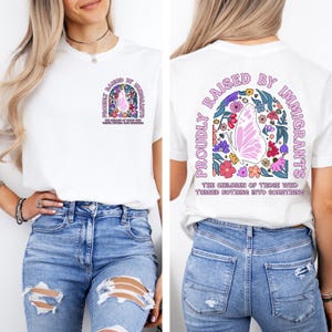 May include: White t-shirt with a colorful graphic on the front and back. The design includes a butterfly, flowers, and the text "PROUDLY RAISED BY IMMIGRANTS" in a decorative arch. The text "THE CHILDREN OF THOSE WHO TURNED NOTHING INTO SOMETHING" is also included.