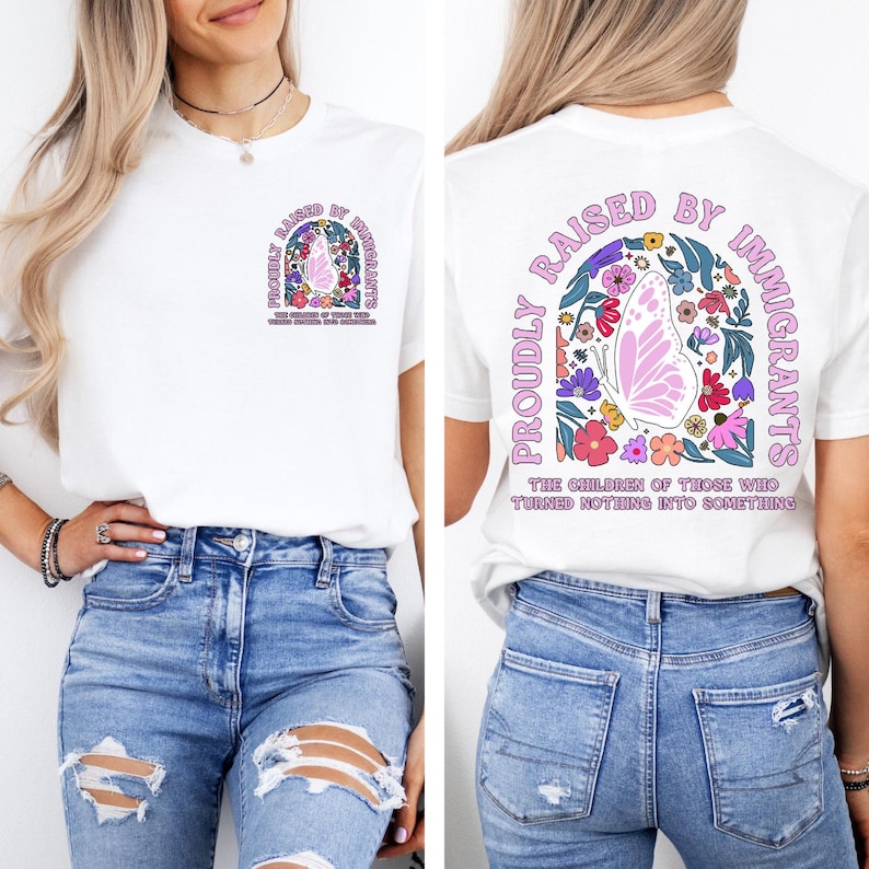 May include: White t-shirt with a colorful graphic on the front and back. The design includes a butterfly, flowers, and the text "PROUDLY RAISED BY IMMIGRANTS" in a decorative arch. The text "THE CHILDREN OF THOSE WHO TURNED NOTHING INTO SOMETHING" is also included.