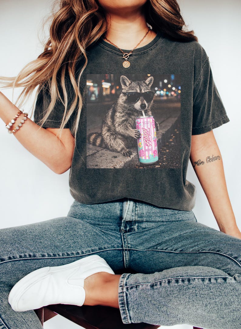 May include: Dark gray t-shirt featuring a graphic of a raccoon wearing sunglasses and holding a pink can with the word "Alani" on it. The raccoon is sitting on a sidewalk with a blurred city background. The shirt is paired with blue jeans.