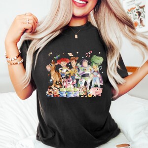 May include: Black t-shirt featuring a colorful graphic of characters from the animated movie Toy Story. The design includes Woody, Buzz Lightyear, Jessie, and other beloved characters. The shirt is a comfortable, casual wear item.