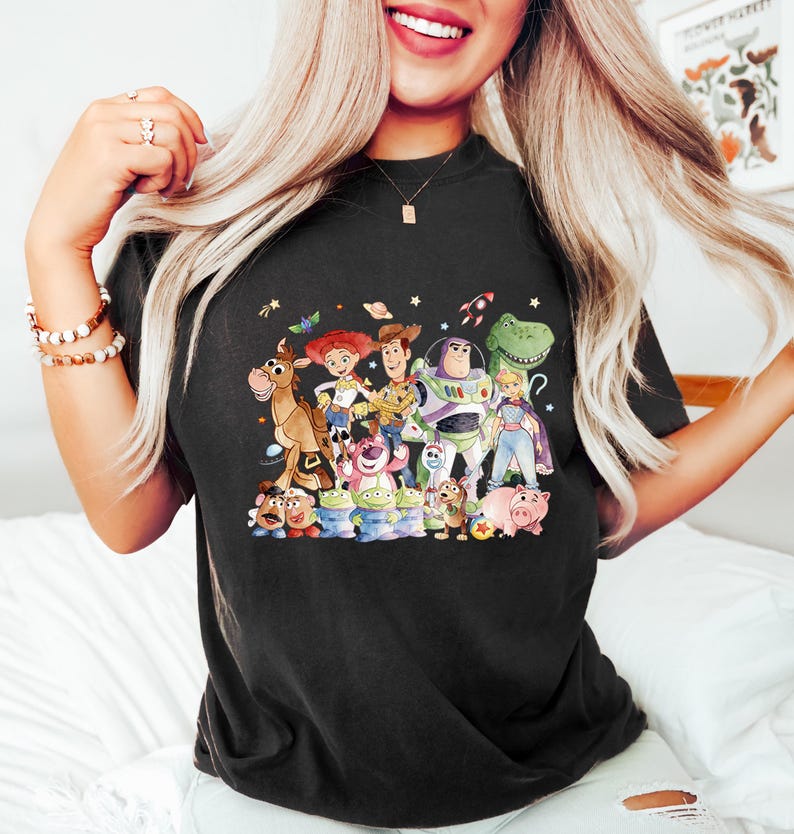 May include: Black t-shirt featuring a colorful graphic of characters from the animated movie Toy Story. The design includes Woody, Buzz Lightyear, Jessie, and other beloved characters. The shirt is a comfortable, casual wear item.