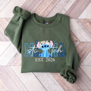 May include: An olive green sweatshirt featuring a blue and white graphic of Stitch with the text "Mama Stitch EST. 2026". The design includes a floral crown and butterflies.